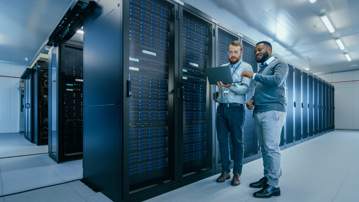 An IT technician wearing glasses and holding a laptop talks to an engineer colleague in a data center.