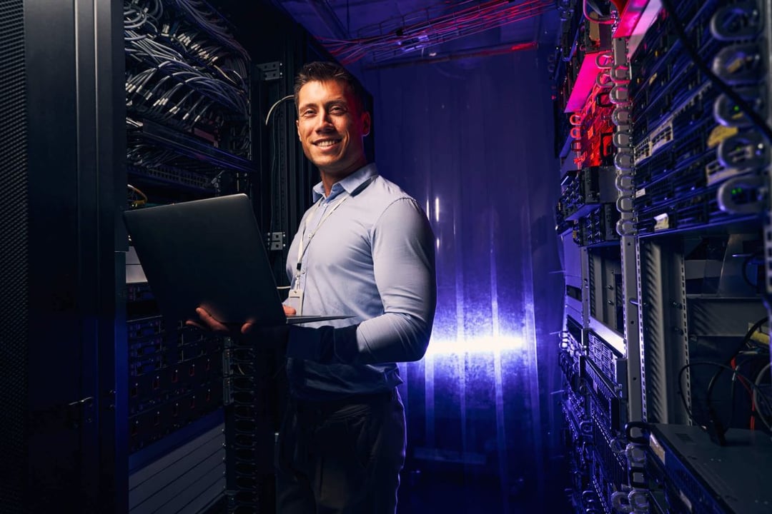 Smiling technician standing in a data center and holding a laptop