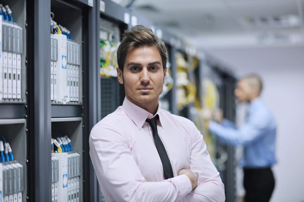 A business person stands in a data center with their arms crossed while a tech works in the background